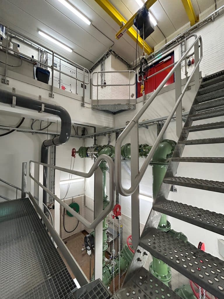 Een totaaloverzicht van de binnenzijde van het rioolgemaal. Bovenin staan de besturingskasten en een takel. Met een wenteltrap kan je naar het deel waar de pompen staan. 